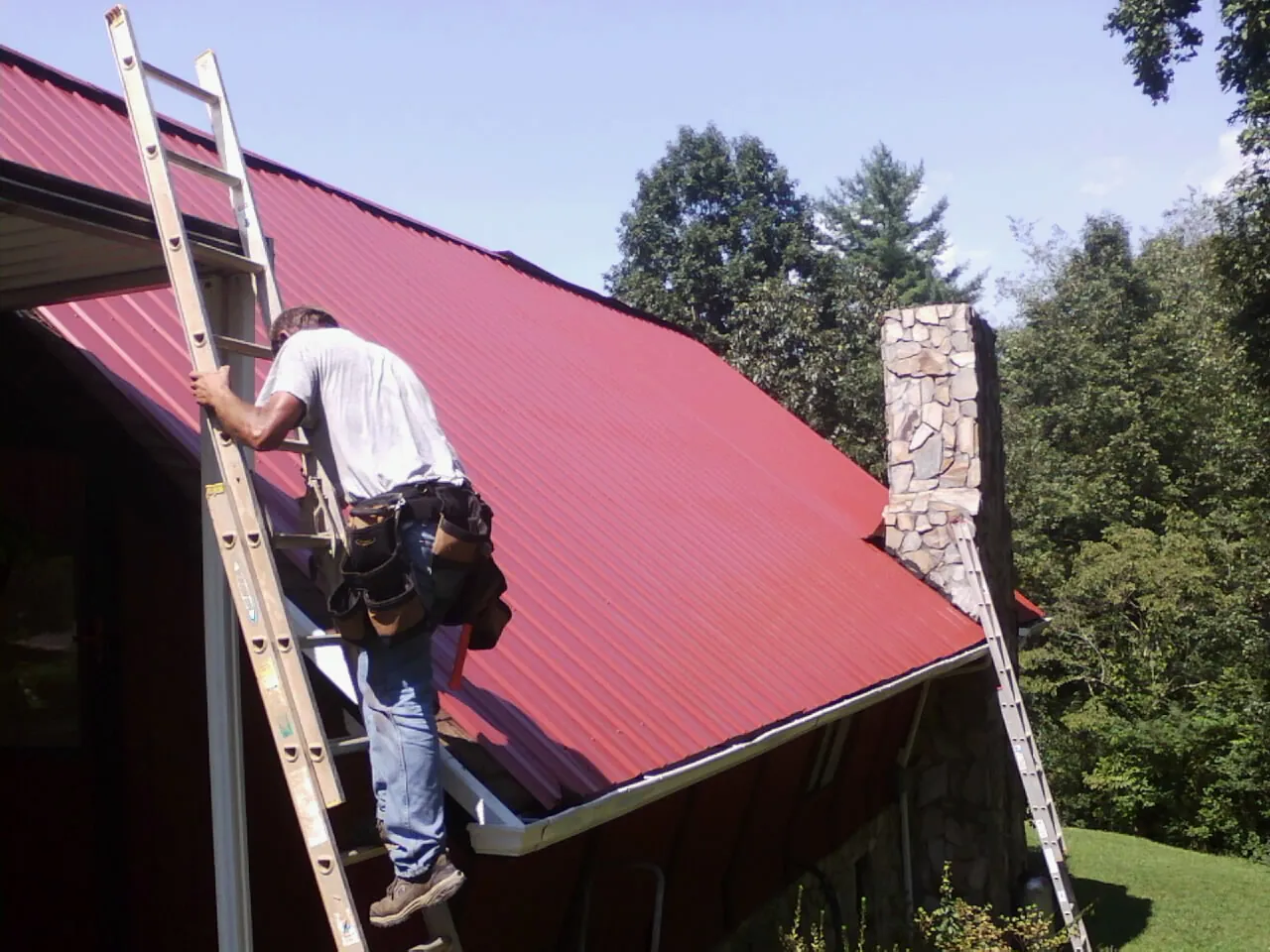 Roofer on a ladder beside a red metal roof and stone chimney — Soffit & Fascia Repair by Roofing Brothers in Snyder